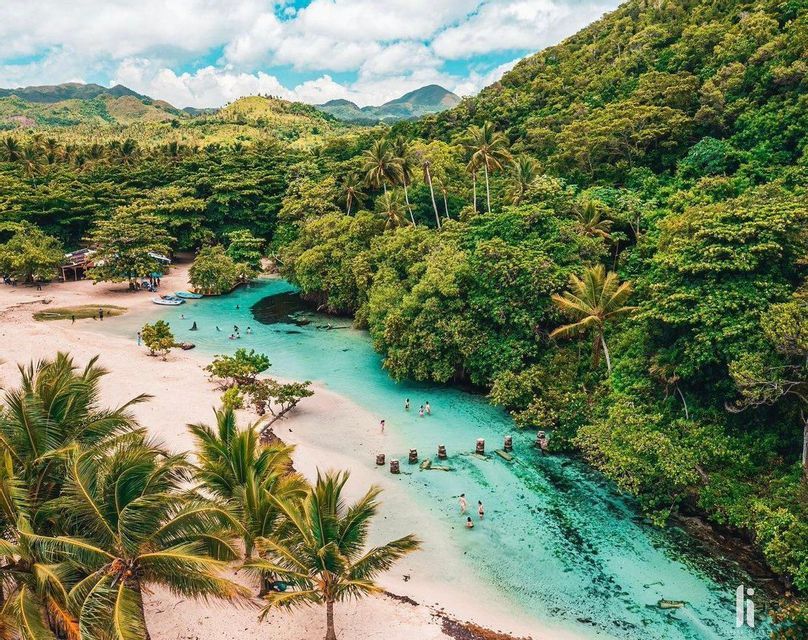 An aerial view of people swimming in the clear turquoise water of a lagoon, edged by a white sand beach and a dense tropical forest.