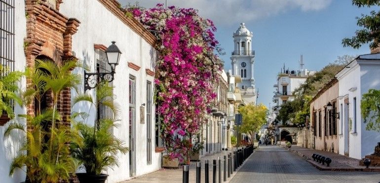 A sunlit cobblestone street lined with white buildings, with pink bougainvillea on a wall and a clock tower in the distance.