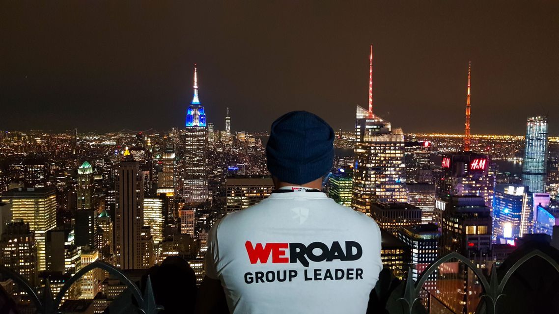 Ein Mann von hinten, der ein 'WeRoad Group Leader'-T-Shirt und eine Beanie trägt und nachts auf eine beleuchtete Stadtkulisse blickt.