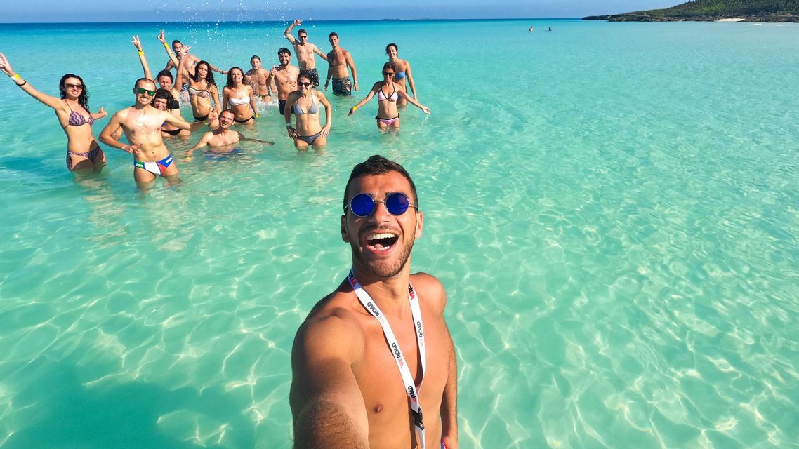 Un homme prend un selfie avec son groupe de voyage WeRoad, tous souriants et debout dans de l'eau turquoise limpide.