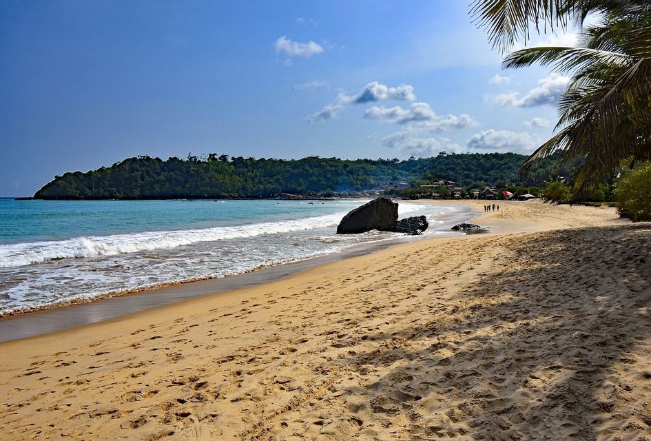 Ein breiter Sandstrand mit Fußspuren führt zu türkisfarbenen Meereswellen, mit einem großen Felsen am Ufer und einem bewaldeten Hügel im Hintergrund.