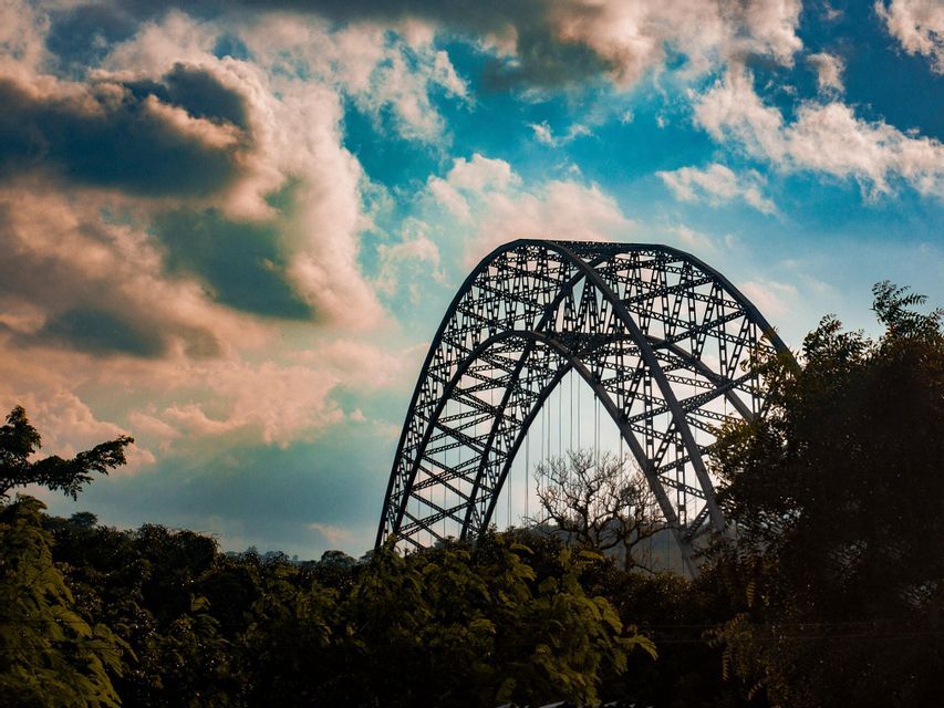 Eine große Stahlgitterbogenbrücke ragt aus einem dunkelgrünen Wald hervor unter einem Himmel mit rosa und blauen Wolken.