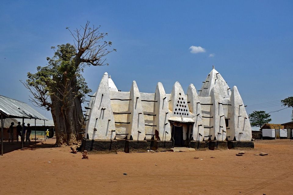 Eine weiße, traditionelle Adobemoschee mit herausragenden Holzbalken steht auf sandigem Boden neben einem großen Baobab-Baum.