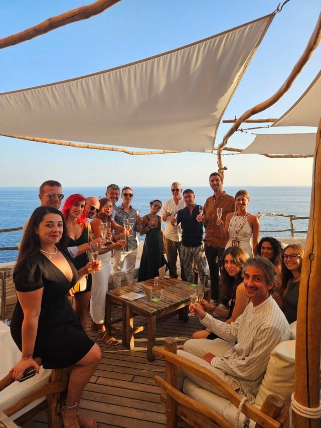 Un viaggio di gruppo WeRoad con persone che brindano con calici di champagne su una terrazza di legno con vista sull'oceano.