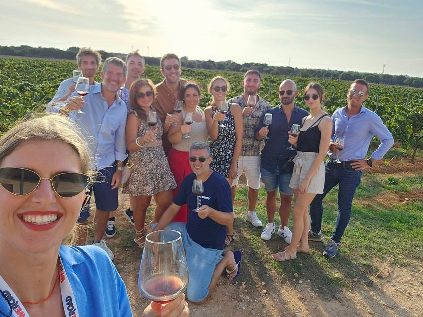A WeRoad group trip posing for a selfie while toasting with glasses of red wine in a sunny vineyard.