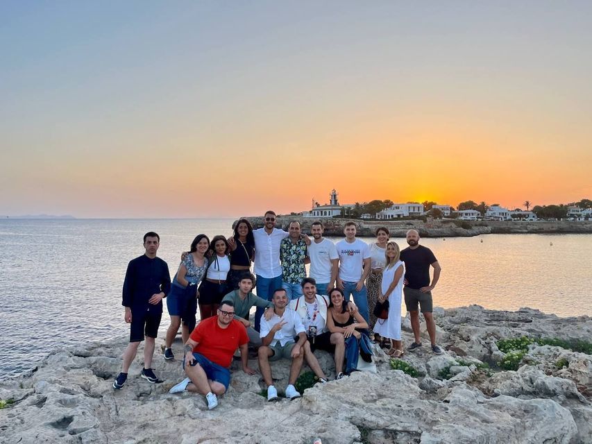 Un gruppo WeRoad in posa per una foto su una costa rocciosa in riva al mare, al tramonto dorato, con un faro in lontananza.