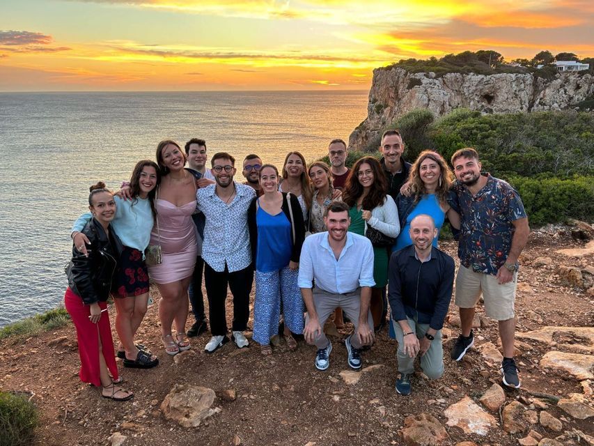 A WeRoad group trip poses for a photo on a coastal cliff with the ocean in the background during a golden sunset.