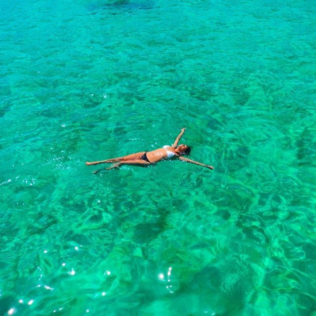 Una donna in bikini fluttua sulla schiena con le braccia aperte in acqua cristallina e turchese, vista dall'alto.