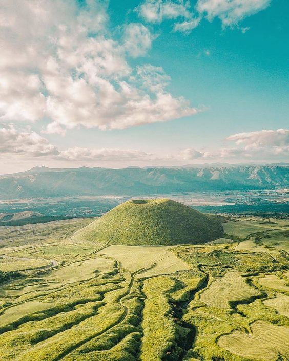 Vista aerea di una collina conica coperta d'erba e con un cratere, circondata da dolci campi verdi sotto un cielo parzialmente nuvoloso.