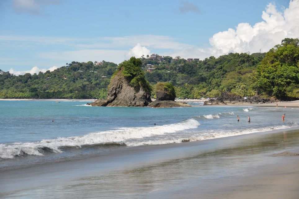 Small waves roll onto a sandy beach where people swim near large rock formations and a lush green hill under a blue sky.