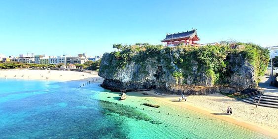 Un tempio giapponese arroccato su una scogliera rocciosa sopra una spiaggia sabbiosa con acque cristalline color turchese e una città sullo sfondo.