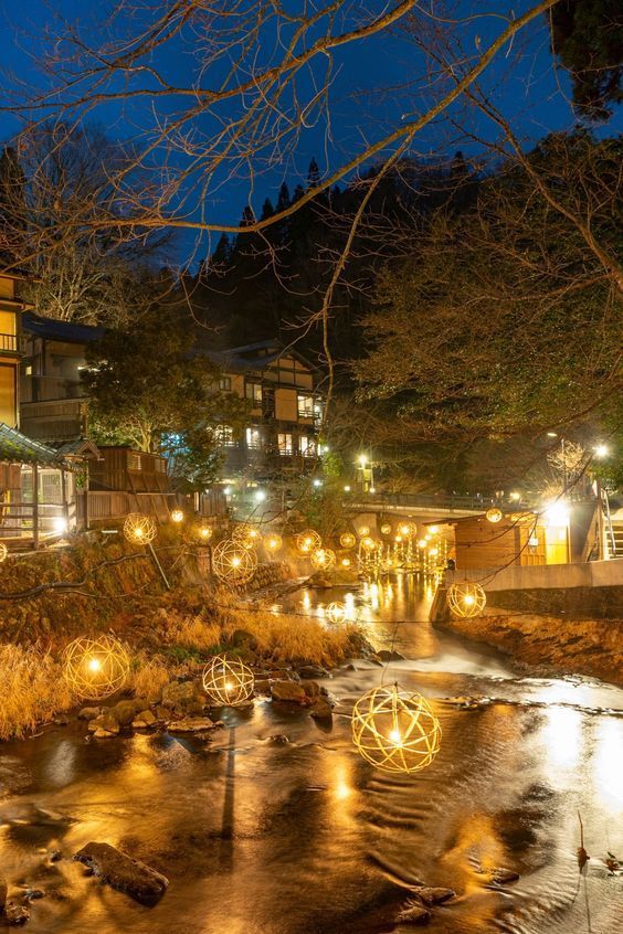 Lanterne sferiche intrecciate illuminano un fiume che costeggia edifici tradizionali di notte.