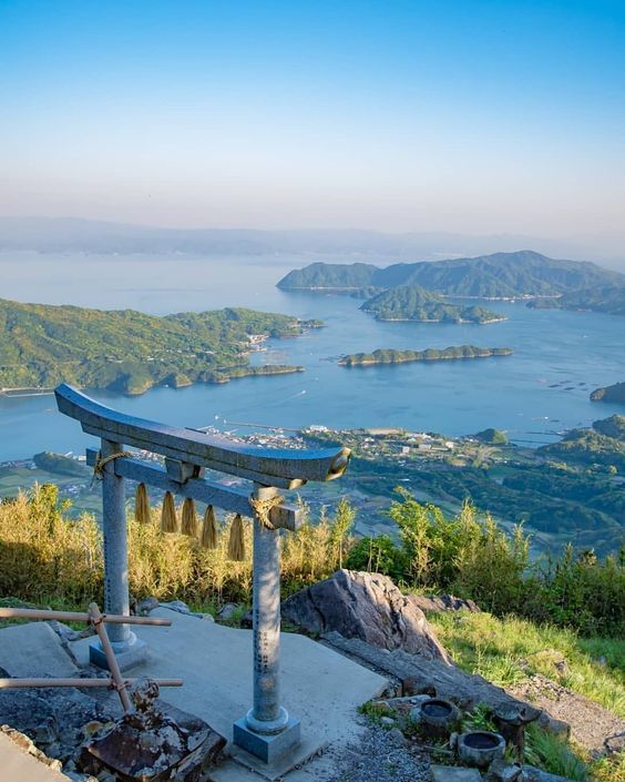 Un torii in pietra sulla vetta di una montagna, affacciato su una baia con numerose isole verdi sotto un cielo azzurro e limpido.