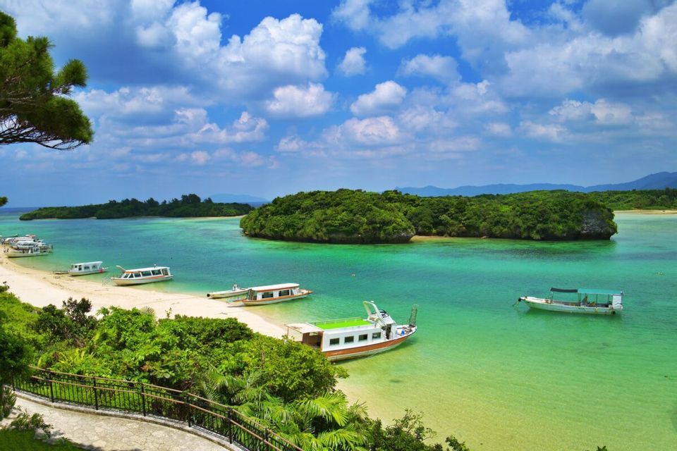 Barche turistiche sono ormeggiate su una spiaggia di sabbia bianca e nell'acqua turchese accanto a un'isola verde lussureggiante sotto un cielo nuvoloso.