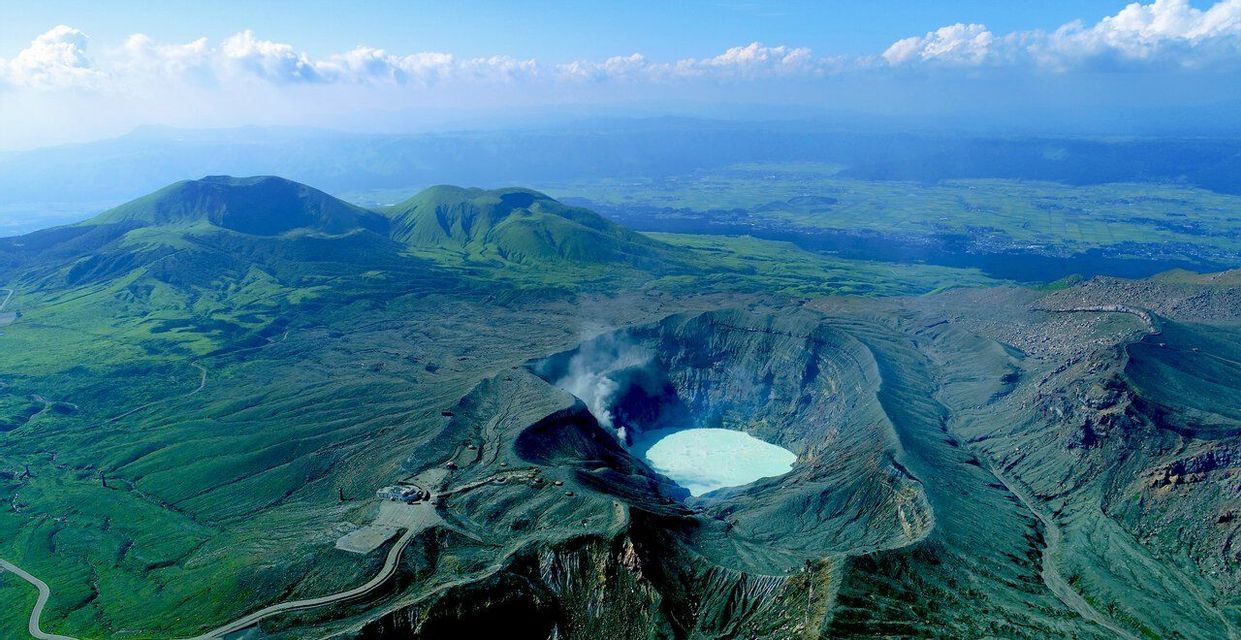 Vista aerea di un cratere vulcanico attivo con un lago turchese, circondato da vaste montagne verdi sotto un cielo limpido.