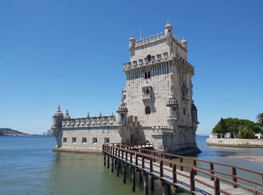 Una fortezza torre in pietra ornata si erge sull'acqua, con una lunga passerella di legno che conduce al suo ingresso in una giornata di sole.