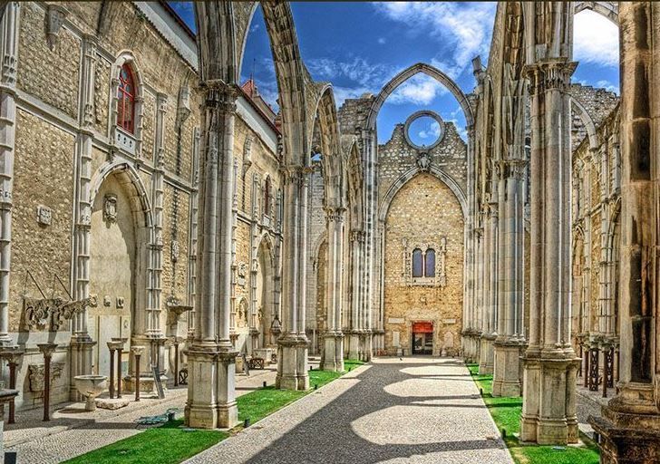 L'interno di una cattedrale gotica senza tetto, con alti pilastri di pietra e archi che proiettano lunghe ombre su una passerella.