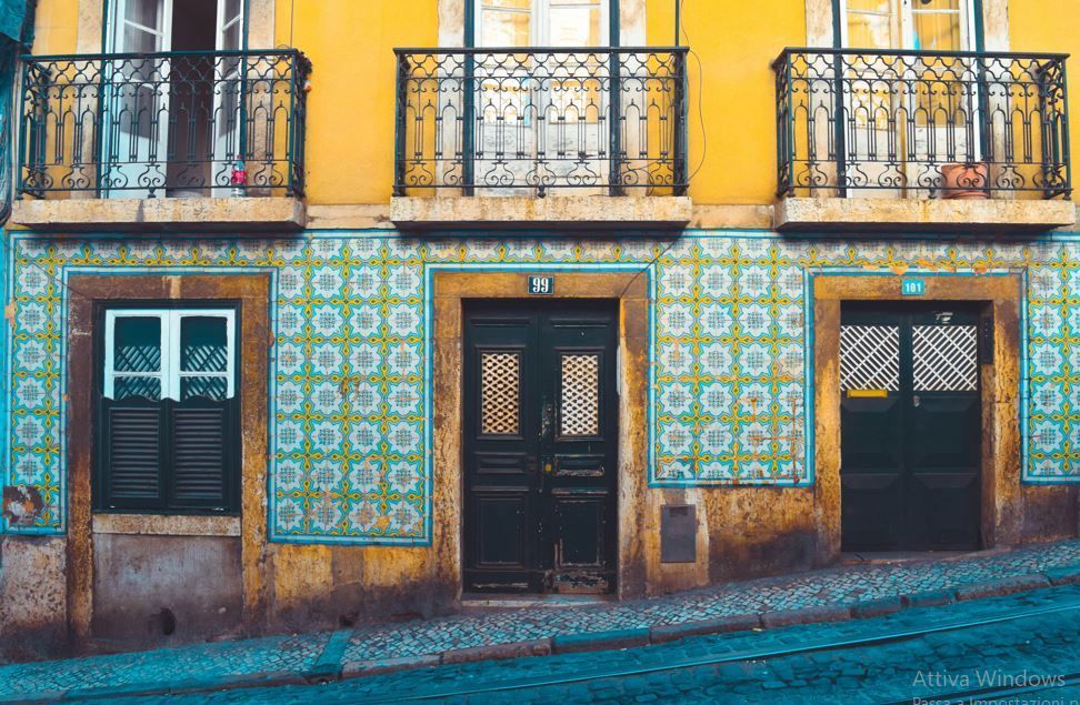 Una facciata gialla di un edificio con piastrelle decorative blu e gialle, balconi neri e porte scure su una ripida strada acciottolata.