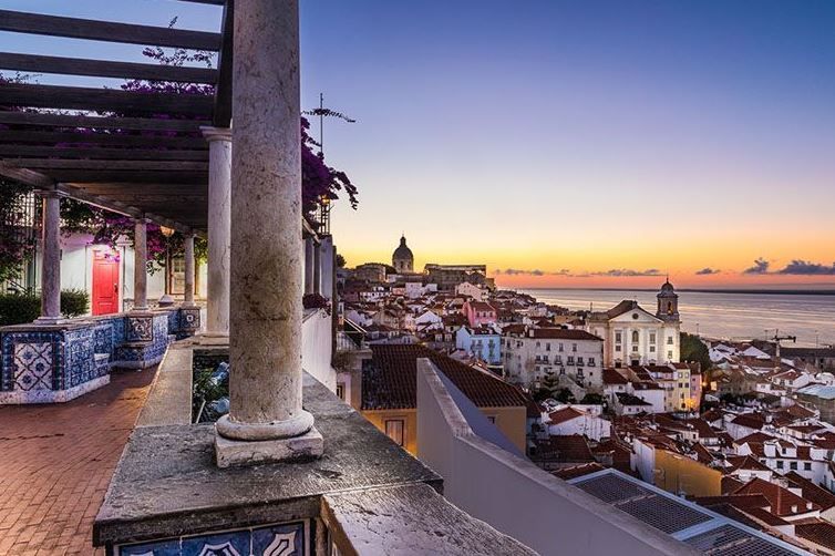 Una vista da una terrazza piastrellata con pilastri in pietra e una pergola che si affaccia su una città costiera con edifici bianchi al tramonto.