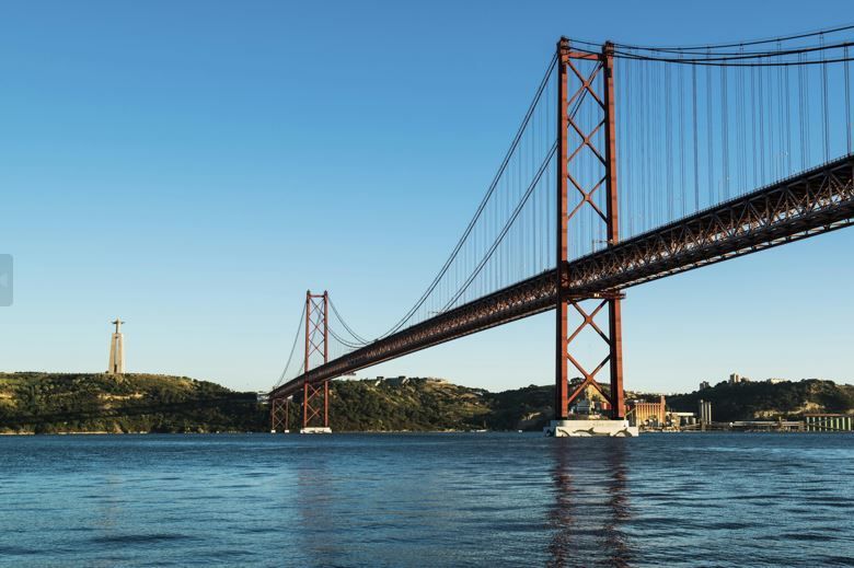 Un grande ponte sospeso rosso attraversa un ampio specchio d'acqua, con un'alta statua su una verde collina distante sotto un cielo azzurro chiaro.