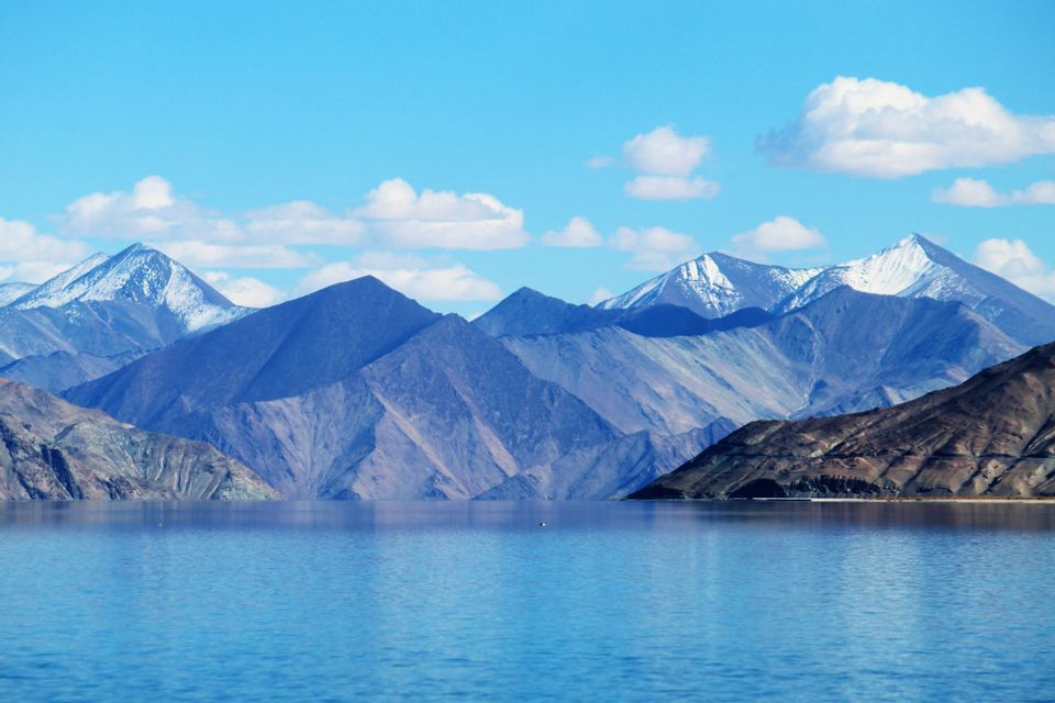 Una catena di montagne rocciose con vette innevate si erge dietro un lago blu calmo sotto un cielo con nuvole bianche.