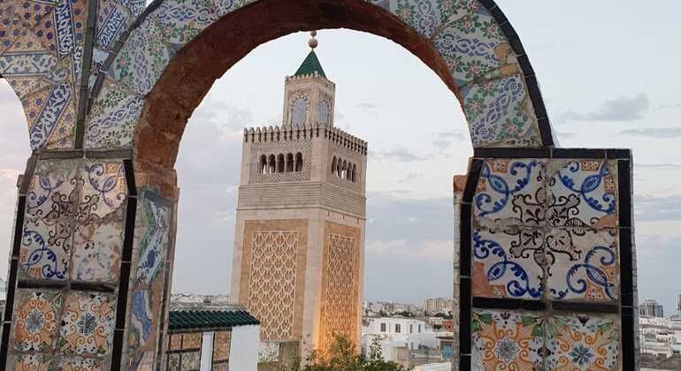 Un minarete de tejado cónico verde visto a través de un arco desgastado, decorado con coloridos mosaicos y con vistas a un paisaje urbano.
