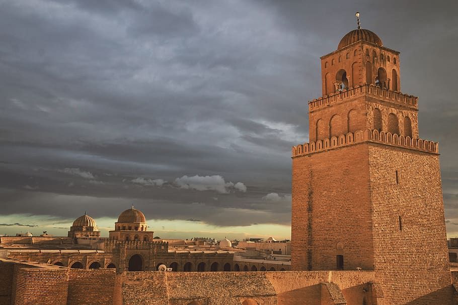 Un minarete de piedra de una mezquita captura la luz dorada del atardecer, con otras cúpulas y tejados visibles contra un cielo nublado.