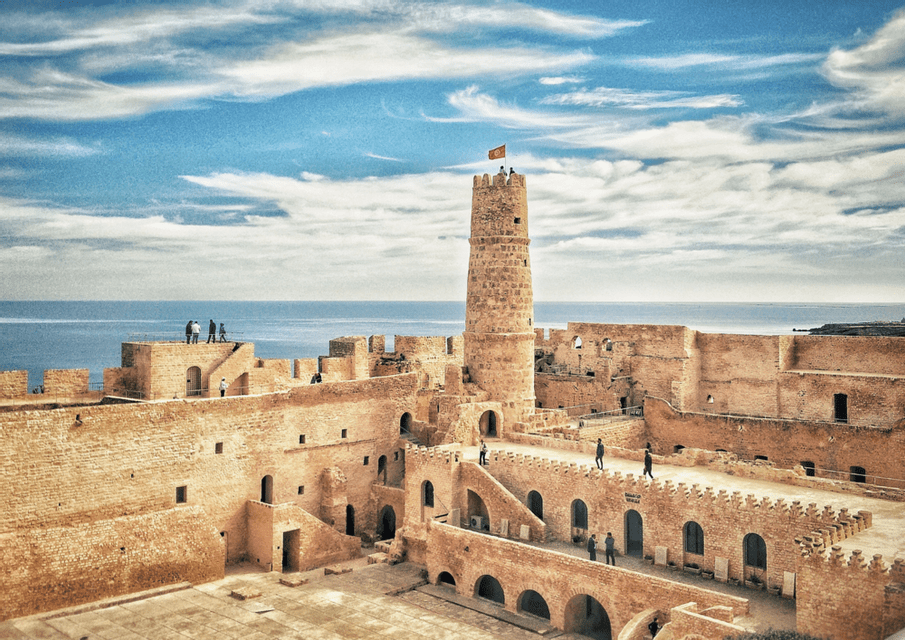 Una fortaleza histórica de piedra con una torre central coronada por una bandera domina el mar, con gente paseando por las murallas.