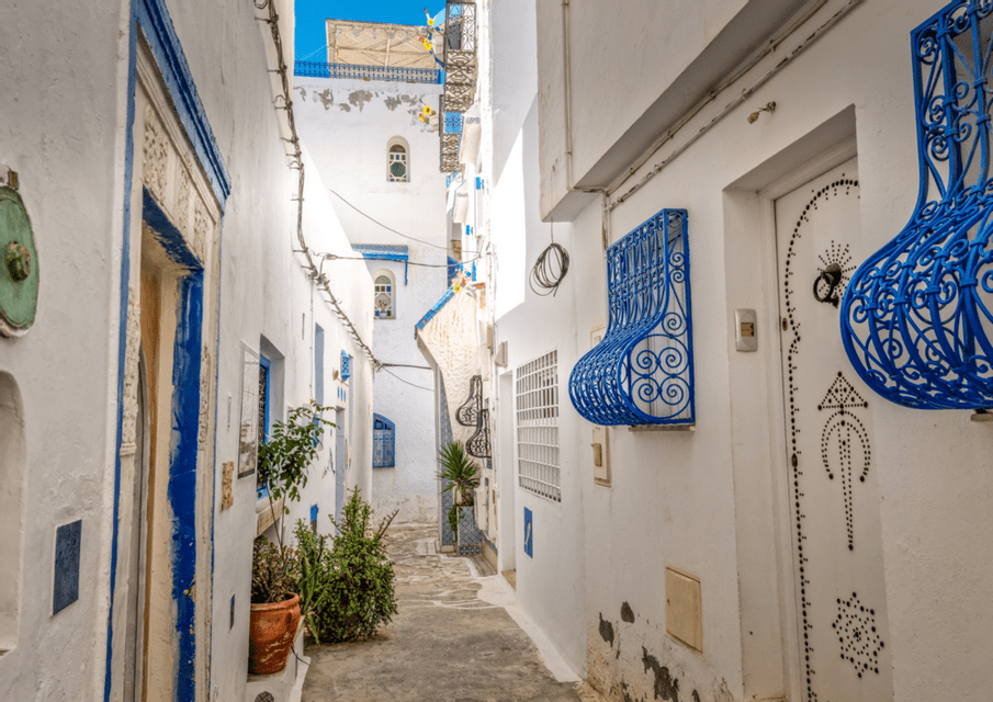Un callejón estrecho con paredes encaladas, puertas azules y rejas de hierro azules ornamentadas bajo un cielo soleado.