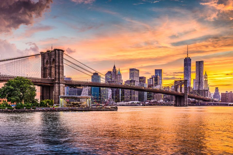 Uno skyline cittadino con grattacieli e un grande ponte sospeso su un fiume, durante un tramonto colorato con nuvole arancioni e viola.