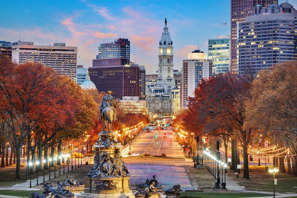 Una statua equestre si affaccia su un ampio viale alberato con alberi autunnali che conduce a uno skyline cittadino con una torre dell'orologio al tramonto.