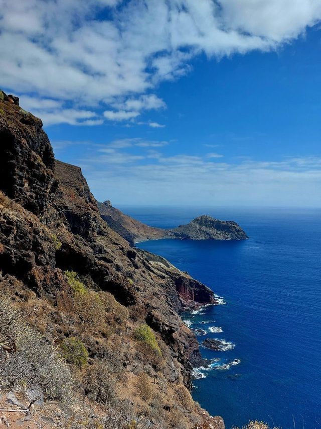 Una vista dall'alto di una ripida costa rocciosa che precipita in un oceano blu intenso sotto un cielo con nuvole bianche.