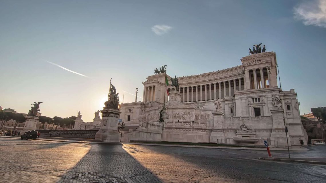 Un grande edificio classico bianco con colonne e statue si erge su una piazza acciottolata all'alba.