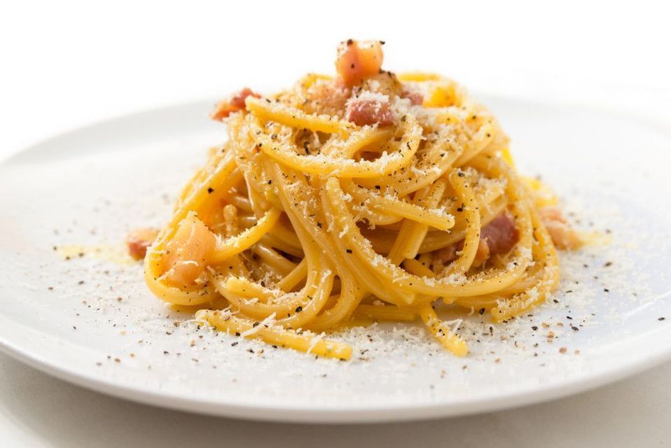 Una porzione di spaghetti alla carbonara su un piatto bianco, guarnita con pancetta, formaggio grattugiato e pepe nero.