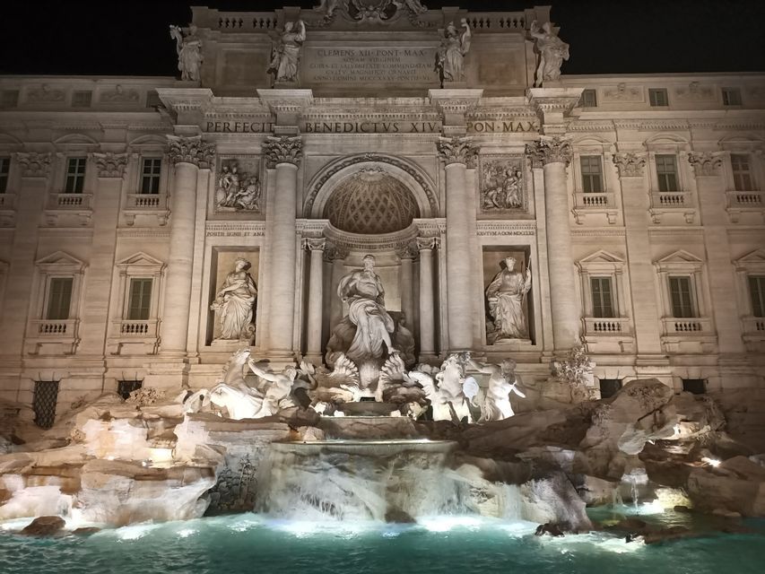 Una grande fontana ornata con sculture classiche, illuminata di fronte a una maestosa facciata di un edificio di notte.