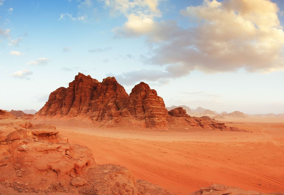 Large red rock formations stand in a vast desert under a partly cloudy sky at dusk.