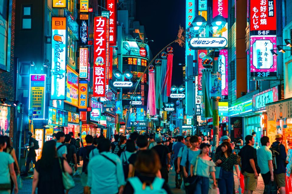 Una gran multitud de personas camina por una estrecha calle de la ciudad por la noche, iluminada por vibrantes letreros de neón multicolores en japonés.
