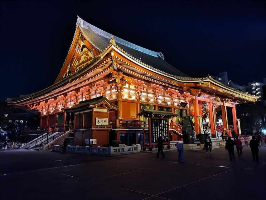An ornate, multi-tiered Japanese temple illuminated with warm lights against a dark night sky, with people walking nearby.