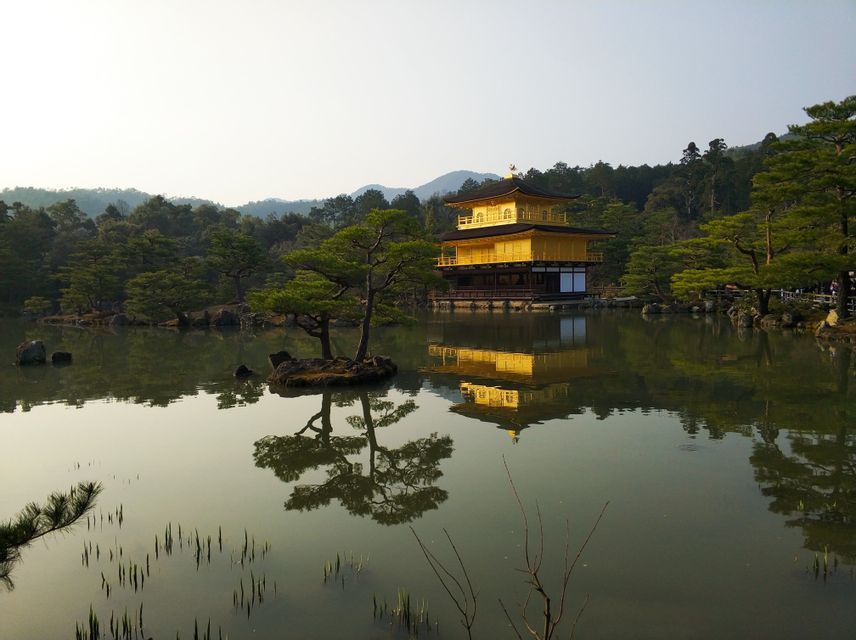 Un temple doré à plusieurs étages se dresse au bord d'un étang, son reflet visible dans l'eau calme près des arbres.