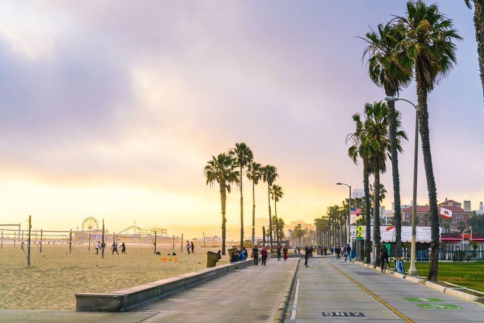Una passerella fiancheggiata da palme, accanto a una spiaggia sabbiosa dove la gente gioca a pallavolo, con un molo divertimenti in lontananza al tramonto.