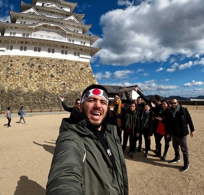 Un homme avec un bandeau japonais prend un selfie souriant lors d'un voyage de groupe WeRoad, devant un château traditionnel japonais.