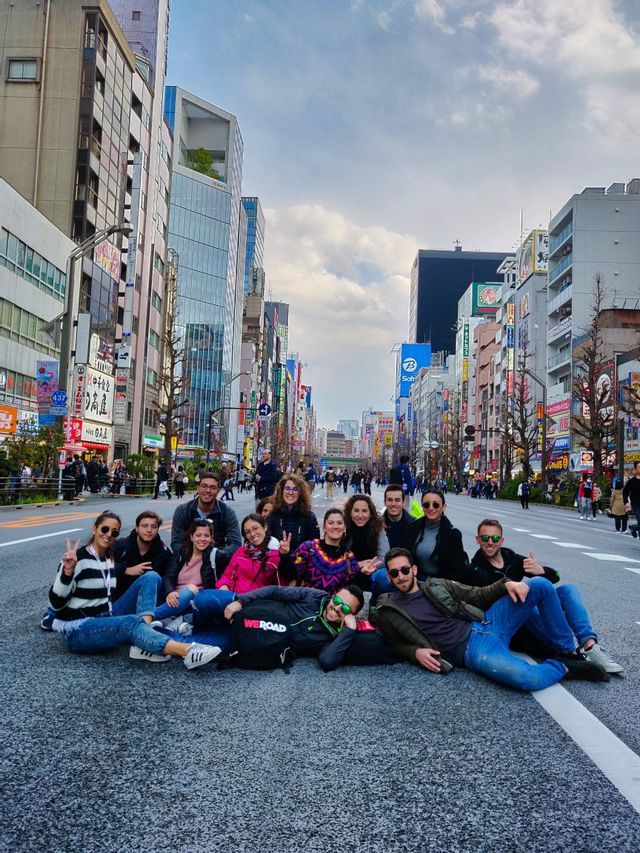 Un grupo de WeRoad se sienta en el centro de una amplia calle de la ciudad, posando para una foto con edificios altos de fondo.