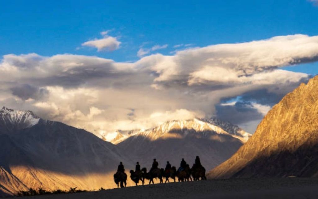 Un viaggio di gruppo WeRoad con sagome a dorso di cammello attraverso un paesaggio desertico con montagne innevate illuminate dal sole sullo sfondo.