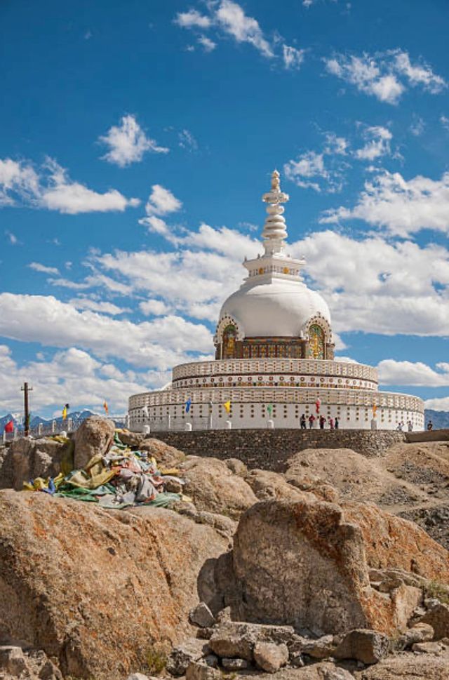 Un grande stupa bianco con una guglia dorata si erge su una collina rocciosa sotto un cielo blu con nuvole bianche sparse.