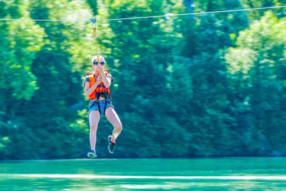 Una donna con giubbotto di salvataggio e imbracatura si lancia in zipline sopra uno specchio d'acqua, con una foresta verde sfocata sullo sfondo.