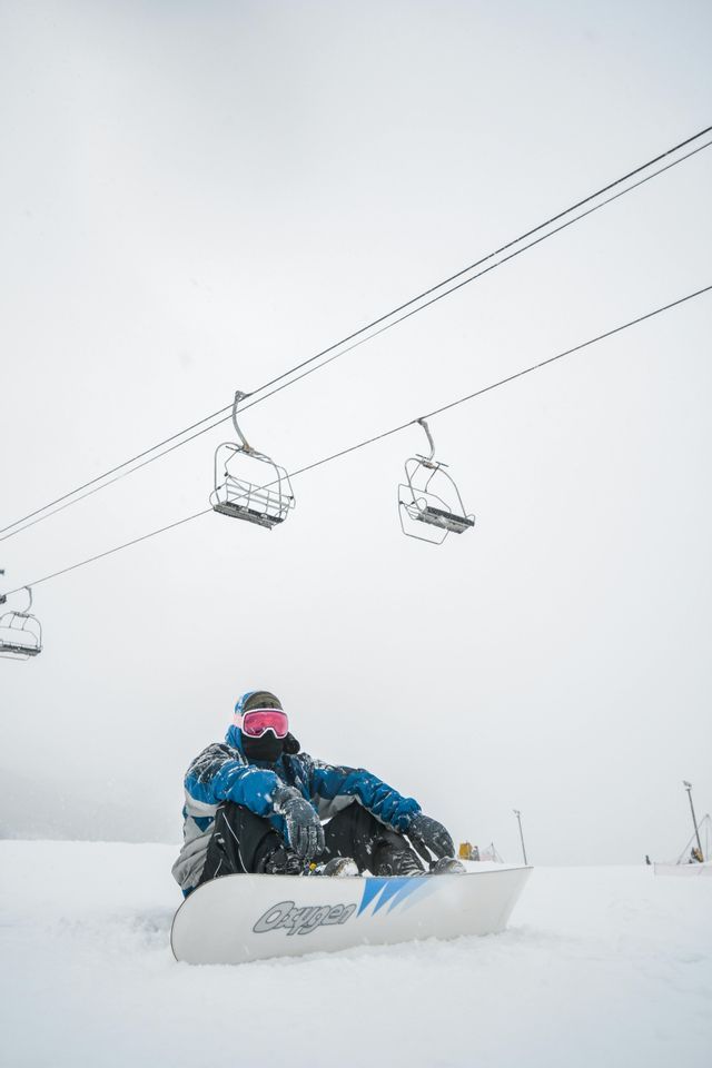 Uno snowboarder con giacca blu siede su un pendio innevato con la sua tavola sotto una seggiovia vuota.