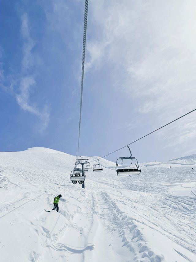 Una persona con una giacca verde fa snowboard su un pendio di montagna, sotto una seggiovia che trasporta un gruppo WeRoad.