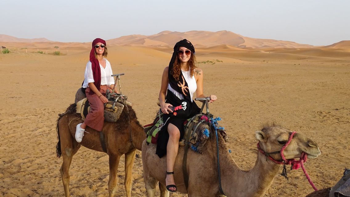 Two women from a WeRoad group trip smiling while riding camels across a vast desert with sand dunes in the background.