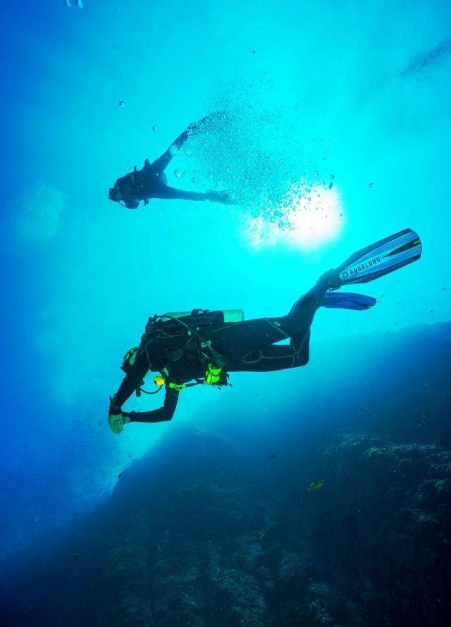Dos buceadores en un viaje grupal de WeRoad nadan en agua azul profunda, con el sol brillando desde la superficie.