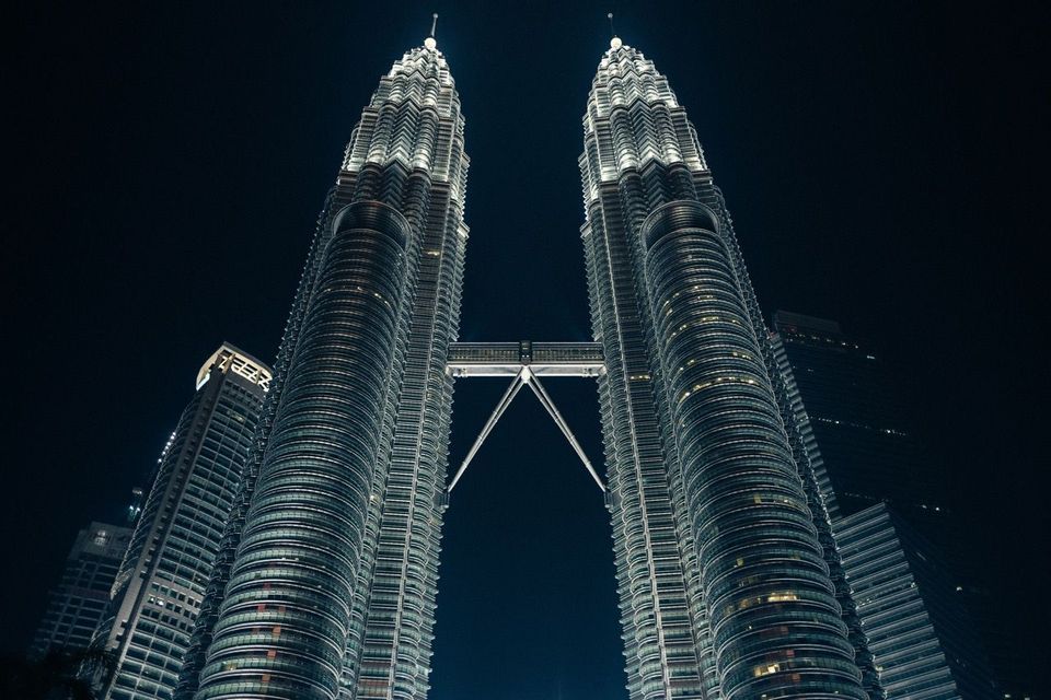 Vista en contrapicado de dos rascacielos idénticos unidos por un puente aéreo, brillantemente iluminados bajo un cielo nocturno oscuro.